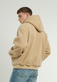 Beige, fleece hettejakke med en teksturert, fluffy overflate. Har en avslappet passform og ribbestrikket kant. Båret over blå denim jeans.