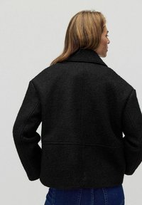 Czarna kurtka wełniana z mieszanki, o pudełkowym kroju, szerokimi klapami i widocznym przeszyciem. Materiał wydaje się teksturowany, prezentując miękkie wykończenie.