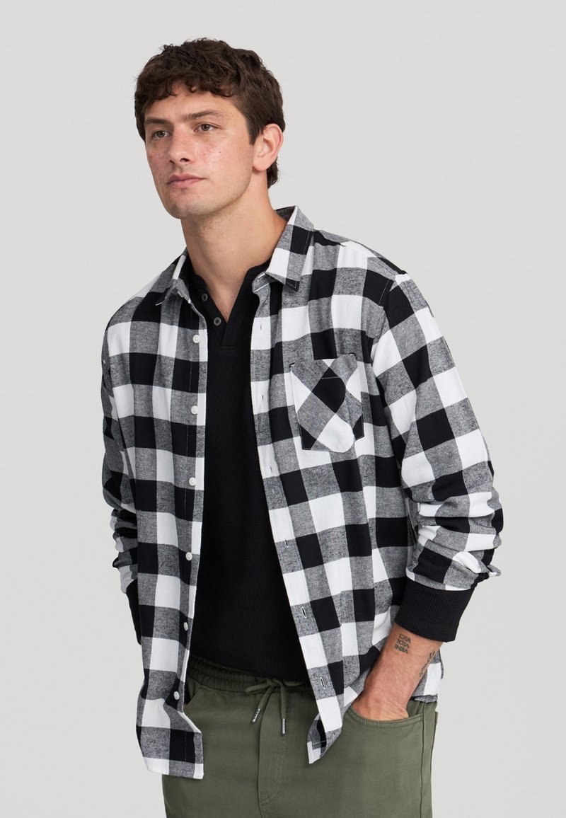 Un giovane con capelli ricci indossa una camicia a scacchi bianco e nero sopra un henley nero, con le mani nelle tasche dei pantaloni verde oliva su uno sfondo bianco.