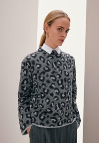 Vrouw met blond haar in een strakke paardenstaart draagt een grijze trui met luipaardprint over een witte blouse en gestreepte broek.