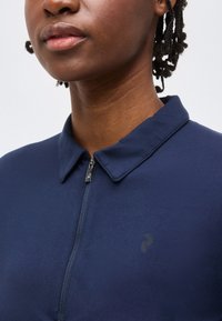 Κοντινό πλάνο ενός ατόμου που φοράει μπλε σκούρο πουκάμισο με φερμουάρ, μυτερό γιακά και διακριτικό λογότυπο στο στήθος.