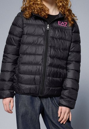 Personne portant une veste matelassée noire EA7 Emporio Armani avec un logo rose sur la poitrine, associée à un jean bleu foncé sur fond gris.