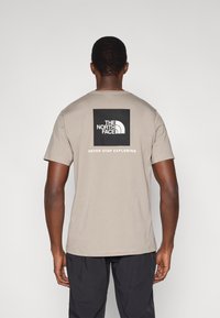 Ανοιχτό μπεζ κοντομάνικο μπλουζάκι με μαύρο τετράγωνο λογότυπο στην πλάτη που γράφει «THE NORTH FACE» και «NEVER STOP EXPLORING.»