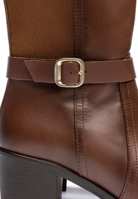 Brauner kniehoher Stiefel aus Leder mit einem Textilpaneel, einem breiten Riemen mit einer quadratischen Metall-Schnalle und einem strukturierten Absatz. Mit Nahtdetails.