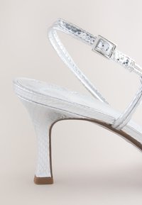 Ezüst színű, kígyóbőr mintás magas sarkú szandál, vékony bokapántokkal és karcsú tűsarokkal. Sima belső talp minimális díszítéssel.