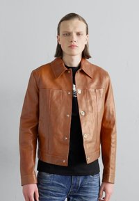 Braune Lederjacke mit Knopfverschluss, spitzem Kragen und zwei Brusttaschen, getragen über einem schwarzen Shirt und Jeans.