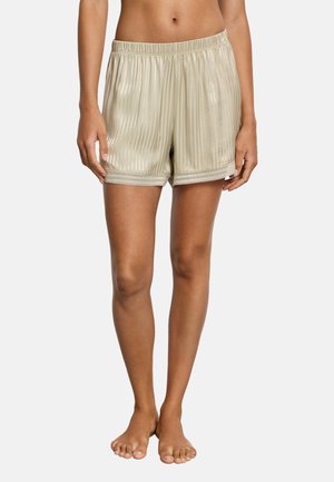 Beige satinshorts med lodrette striber og blonderkant båret af en person, der står barfodet mod en hvid baggrund.