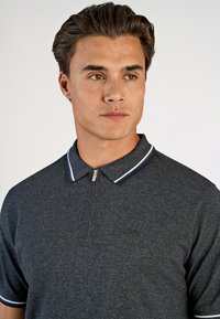 Mörkgrå polo skjorta med dragkedja i kragen, med ett vitt och marinblått randigt accent vid kragen. Mjukt, texturerat tyg med subtila detaljer.