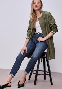 Mujer sentada en un taburete negro, vistiendo una chaqueta bomber verde oliva, camiseta blanca sin mangas, jeans azules remangados y zapatos planos negros con punta.