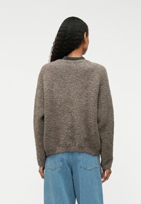 Pulover texturat de culoare maronie, cu guler rotund, mâneci lungi și croială lejeră, purtat deasupra unui blug albastru cu talie înaltă.