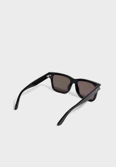 Lunettes de soleil rectangulaires noires avec une monture épaisse et brillante et des verres foncés, présentées légèrement inclinées sur un fond gris clair.