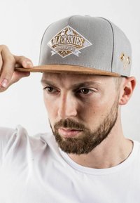 Gorra de béisbol gris con visera de cuero beige, con un logo bordado en dorado y blanco. El diseño incluye hachas cruzadas como acentos.
