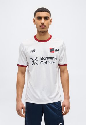 Joven con camiseta blanca de fútbol con ribete rojo, logotipo de Barmenia Gothaer y emblema 04 Leverkusen, de pie frente a un fondo liso.