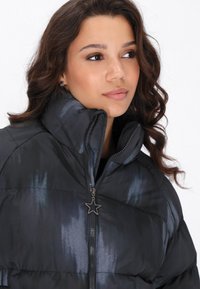Zwarte pufferjack met glans, hoge kraag en rits met een stervormige trekker. Gelaagd stof met subtiele donkblauwe patronen.