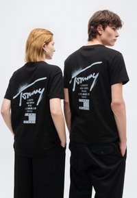 Δύο άτομα που φορούν μαύρα t-shirt Tommy Jeans με τυπωμένα ονόματα πόλεων στην πλάτη, στέκονται δίπλα δίπλα μπροστά σε λευκό φόντο.