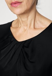 Černá halenka s jemným, splývavým výstřihem a řasenými detaily na přední straně. Tkanina působí hladce a lehce, s přiléhavým střihem.