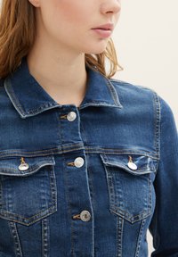 Dark blue denim jacket with a collar, silver buttons, two chest pockets, and visible stitching details. Texture is smooth with a slightly worn appearance.