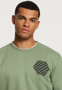 Felpa verde chiaro realizzata in un tessuto morbido, con un motivo esagonale nero testurizzato sul petto. Design a girocollo con colletto a costine.