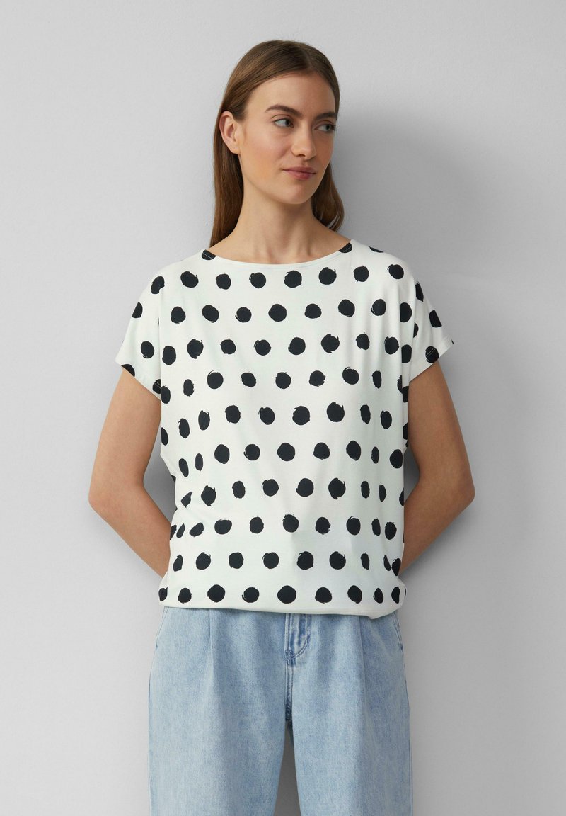 Witte T-shirt met korte mouwen, voorzien van een zwart stippenpatroon, gemaakt van zachte stof met een ontspannen pasvorm en ronde halslijn. Gepaard met lichtblauwe jeans.