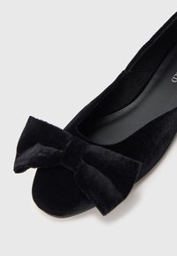 Μαύρο βελούδινο παπούτσι με επίπεδη σόλα και μια μεγάλη βελούδινη κορδέλα στη στρογγυλή μύτη, με λείο μαύρο εσωτερικό, εμφανιζόμενο σε λευκό φόντο.