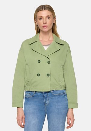 Vrouw draagt lichtgroene dubbelzijdige korte jas met brede kraag en blauwe spijkerbroek, staand tegen een effen achtergrond.