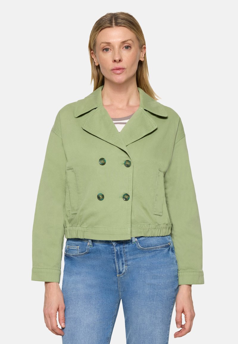 Femme portant une veste courte croisée vert clair avec un large col et un jean en denim bleu, debout devant un fond uni.