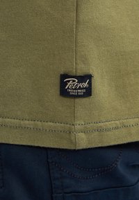 Olivengrøn bomulds T-shirt med en sort etikette, der står "Petrol Industries Since 1989", syet i sømmen, parret med mørk denim. 