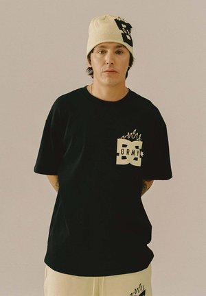 Zwart katoenen t-shirt met een grafische logo in witte en gele vlammen op de linkerkant van de borst, gecombineerd met een crèmekleurige beanie met een letterontwerp.