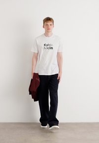 Hvit t-skjorte med rund hals og "Calvin Klein" logo, svarte bukser, og en bordeauxfarget hettegenser holdt i venstre hånd. Hvite sneakers.