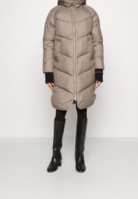 Kvinde iført knælang, taupe pufferjakke med frontlynlås, sorte leggings, sorte knæhøje læderstøvler og sorte strikkede fingerløse handsker.