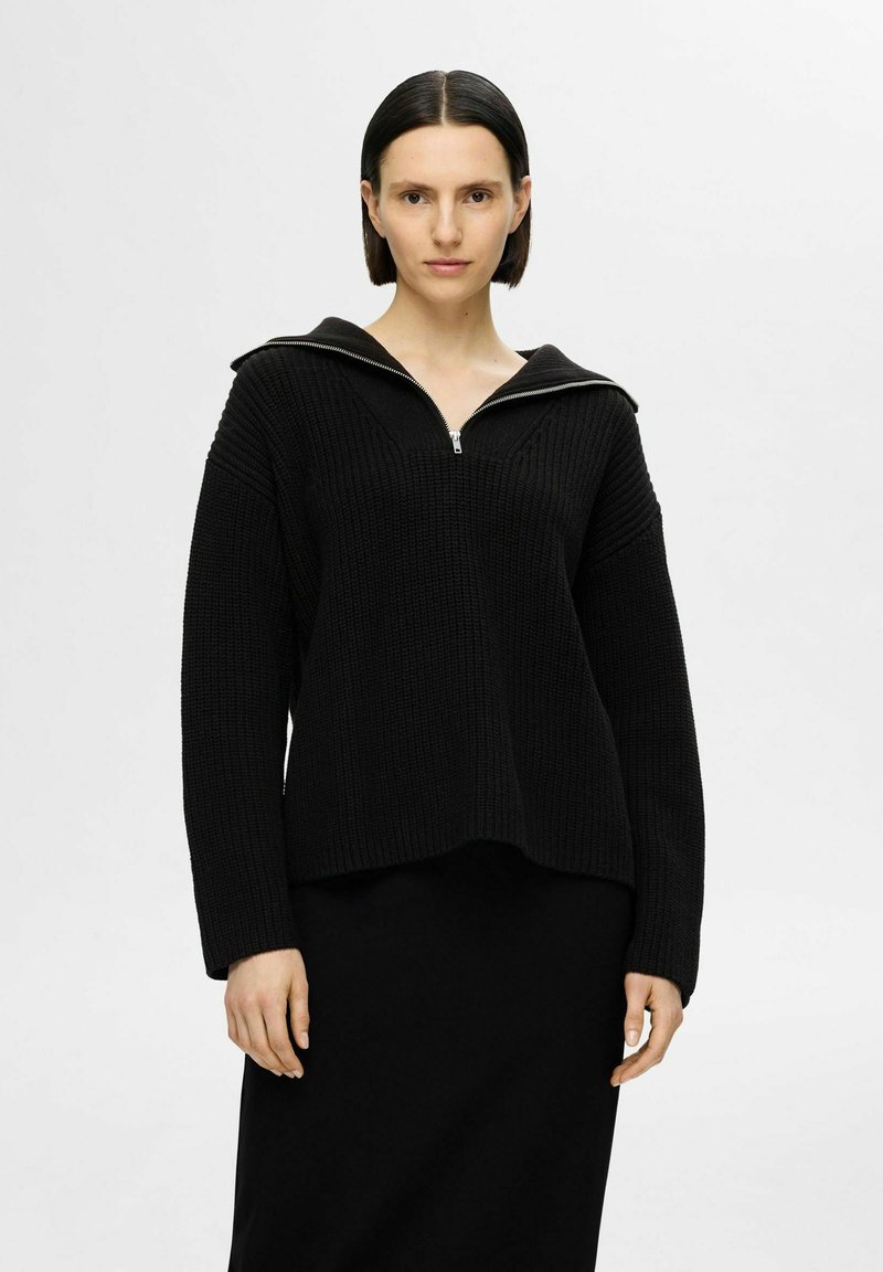 Pullover a zip in maglia nera con un grande colletto strutturato. Tessuto testurizzato, vestibilità ampia e maniche lunghe. Indossato sopra una gonna nera abbinata.