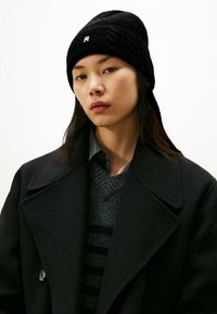 Gorro de punto negro con textura acanalada, que presenta un pequeño logo. Combinado con un abrigo oscuro y un suéter gris a rayas.