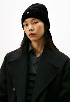 Black knitted beanie with ribbed texture, featuring a small logo. Paired with a dark coat and a gray sweater with stripes.