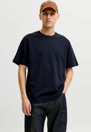 Giovane uomo con cappello marrone, maglietta blu navy e jeans scuri, in piedi con una mano in tasca davanti a uno sfondo semplice.