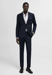 Námornícky dvojdielny oblek so slim fit, klopovými goliermi a bielou košeľou. Čierne kožené topánky dopĺňajú formálny vzhľad.