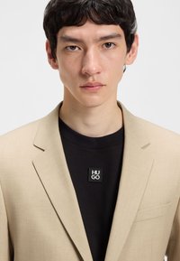 Young man with short dark hair wearing a beige blazer over a black shirt with a small square "HUGO" logo patch.