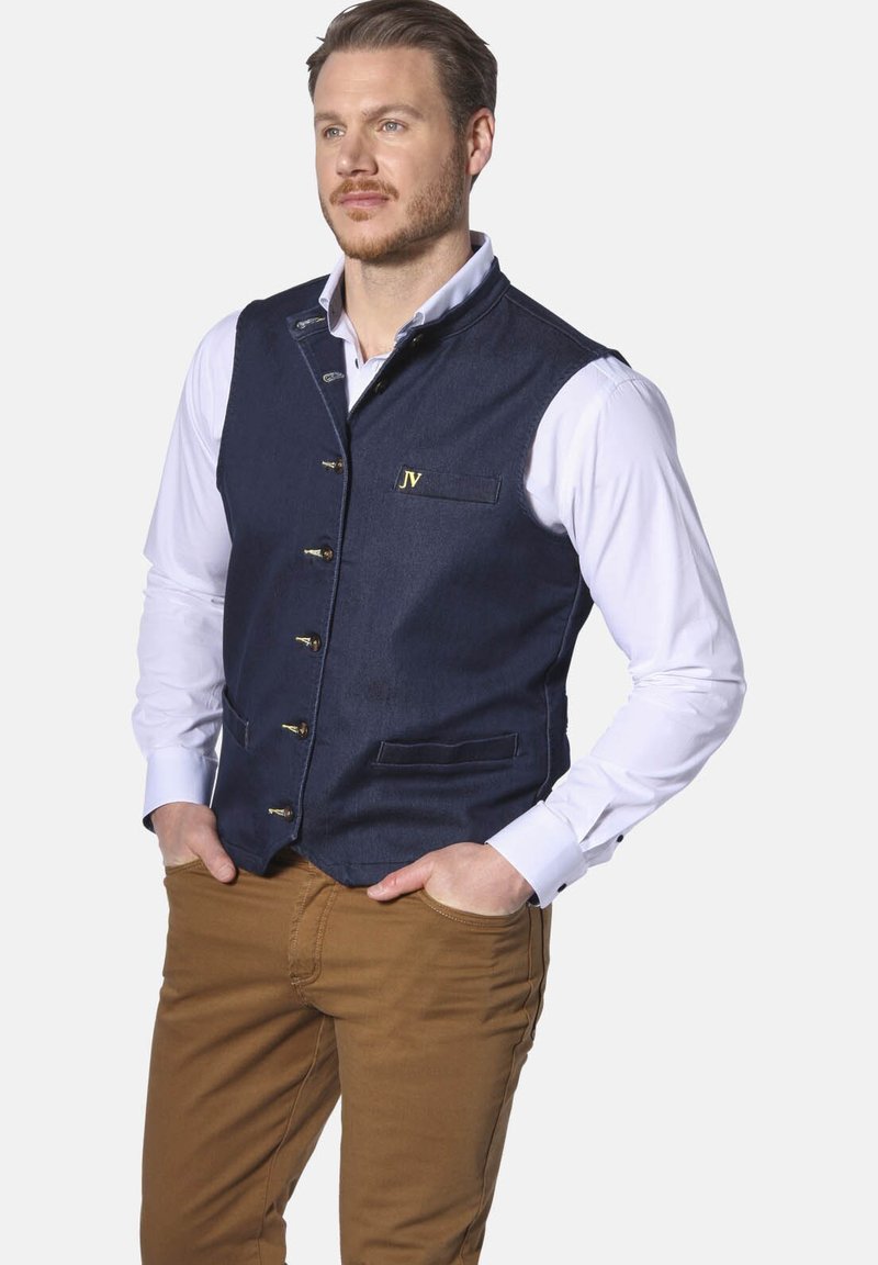 Marineblaues Denim-Weste mit hohem Kragen, goldenen Beschlägen, vorderen Taschen und gesticktem Logo. Über einem weißen Hemd getragen und mit braunen Hosen kombiniert.