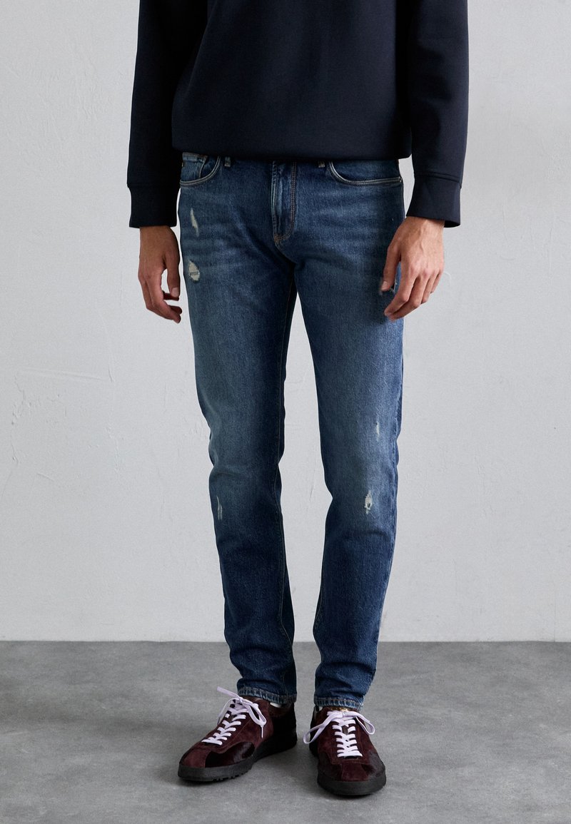 Persona vestida con jeans azules desgastados, zapatillas de deporte burdeos oscuras con cordones blancos y una camiseta negra de manga larga, de pie sobre un suelo gris.