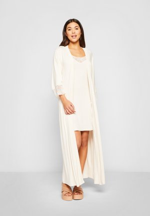 Langer cremefarbener Bademantel mit Spitzenakzenten, kombiniert mit einem figurbetonten cremefarbenen Kleid und klobigen beige Plattformsandalen. Glatter Stoff, fließendes Design.
