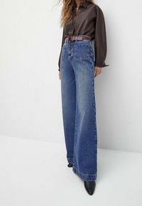 Jean bleu à jambes larges en denim, avec poches avant et fermeture à boutons, associé à une chemise sombre à boutons et une ceinture marron.