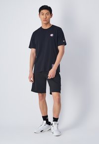 T-shirt in cotone nero con logo sul lato sinistro del petto, abbinata a pantaloni corti neri. Il modello indossa scarpe da ginnastica bianche e calze nere. Design semplice e informale.