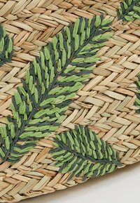 Surface en paille tissée avec des motifs de feuilles brodées en vert foncé et clair, superposés en diagonale.
