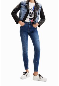 Jeans i denim med färgglada applikationer, tillsammans med en ärmlös jacka i svart och blått och en grafisk t-shirt. Vita sneakers fulländar looken.