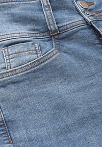 Ljust blå denimtyg med en slät textur, med en framficka, orange sömmar och metallknappdetaljer vid midjan.