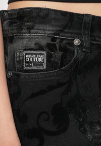 Črne kavbojke iz jeansa z teksturiranim cvetličnim vzorcem. Vključujejo znamko z napisom "VERSACE JEANS COUTURE" in srebrno zaponko.