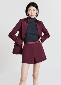 Femme aux cheveux courts noirs portant un blazer bordeaux et un short assorti à un col roulé noir à rayures, se tenant les mains sur les hanches.