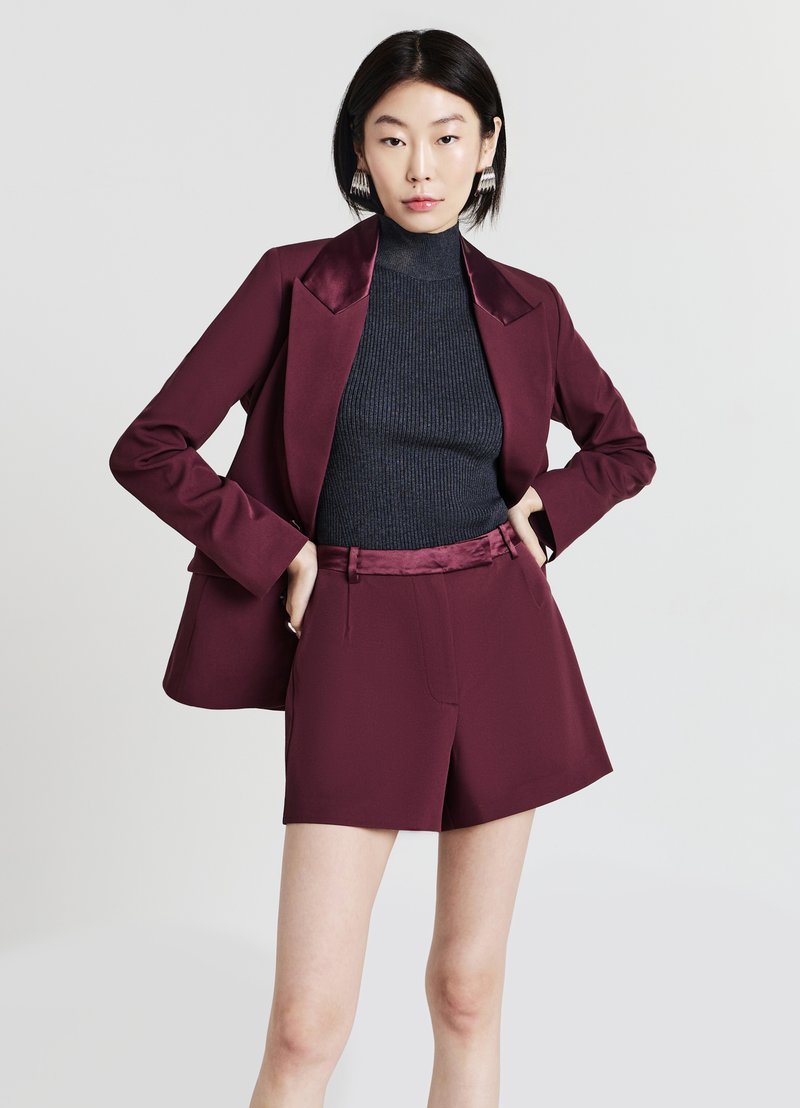 Femme aux cheveux courts noirs portant un blazer bordeaux et un short assorti à un col roulé noir à rayures, se tenant les mains sur les hanches.