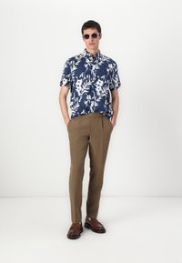 Camisa de manga corta azul marino con estampado floral blanco, combinada con pantalones marrones a medida y mocasines marrones. Modelo con gafas de sol.