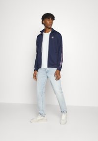 Giacca zip-up blu navy con righe rosse e bianche a contrasto sulle maniche, indossata sopra una t-shirt bianca, abbinata a jeans in denim chiaro e sneaker.