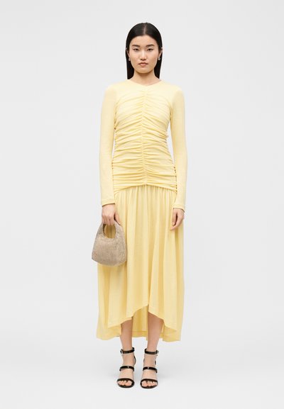 Ženska v rumeno obleko z dolgimi rokavi, ki drži bež teksturirano torbico, obuta v črne sandale z trakci, stoji pred enobarvnim ozadjem.